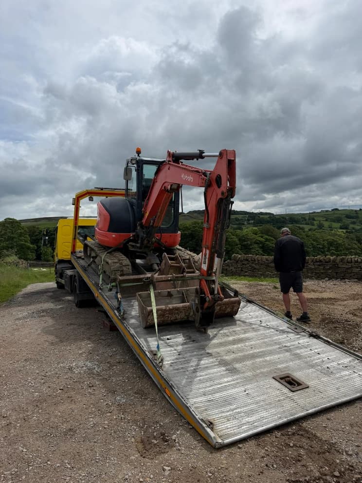 Mini excavator loaded on a flatbed recovery trailer in a rural, hilly location